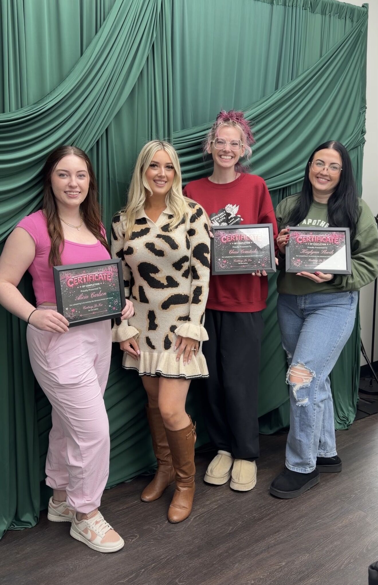 Four graduates holding their De Voe Lashes certificates in front of a green drape
